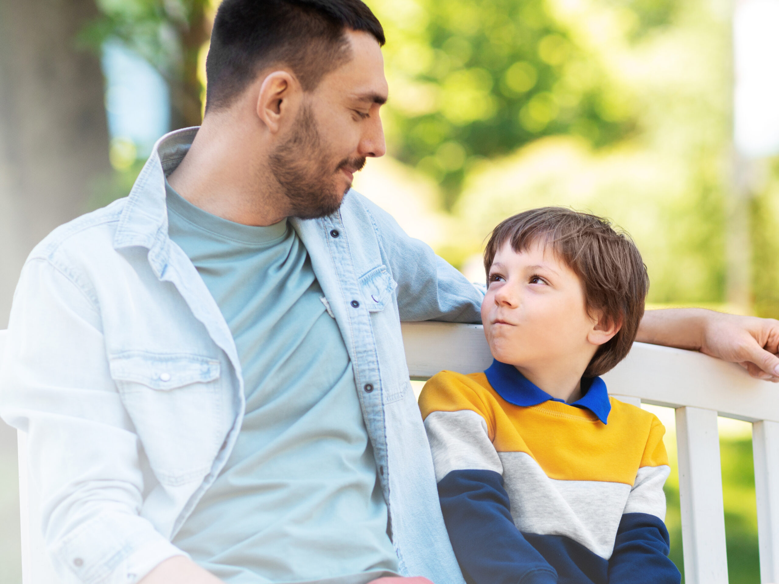 family, fatherhood and people concept - happy father and son sitting on bench at summer park and talking